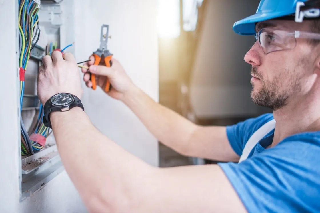 Electrician in blue shirt installing wiring in an open electrical panel with pliers