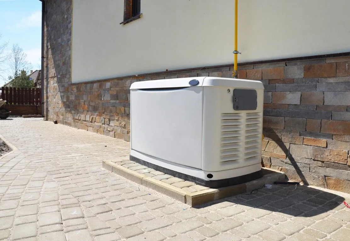Portable generator beside a house wall on a paved driveway, with a yellow pipe and brick foundation.