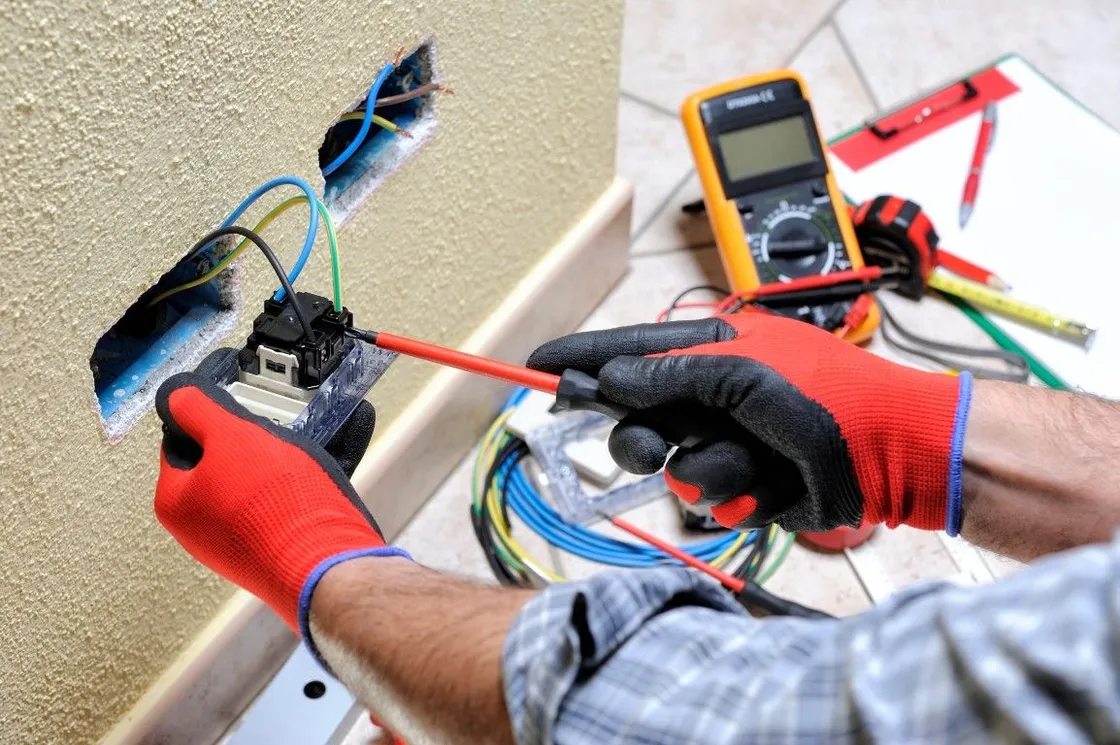 Electrician in red gloves testing wall wiring with a multimeter and tools on a workbench.